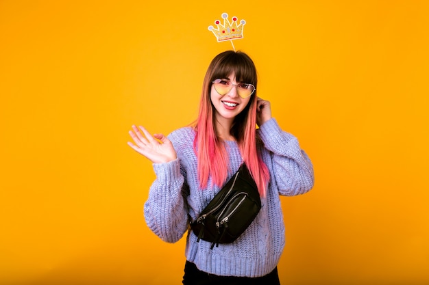 Retrato engraçado brilhante de mulher alegre hippie com cabelo rosa brilhante, vestindo uma camisola confortável, segurando a coroa falsa festa e sorrindo, pronto para a festa, parede amarela.