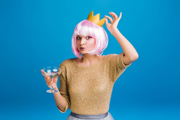 Retrato engraçado elegante jovem de suéter dourado, corte de cabelo rosa, coroa na cabeça. Se divertindo, bebendo champanhe, comemorando festa de ano novo, aniversário.