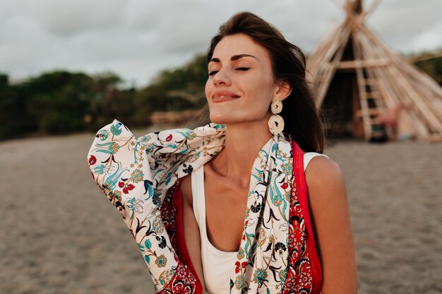 Retrato externo de uma adorável senhora romântica com os olhos fechados, usando um xale da moda e ganhos elegantes descansando na praia em um dia ensolarado de verão