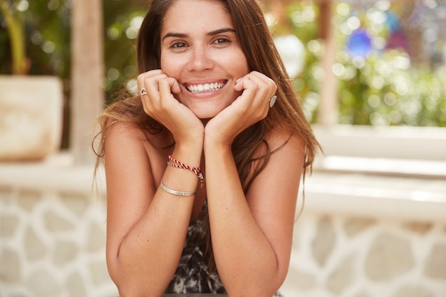 Retrato horizontal da modelo feminina alegre e encantada com sorriso largo mantém as mãos embaixo do queixo, recrie em um café, descansa no verão em uma ilha tropical