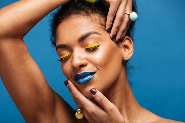 Retrato horizontal da mulher afro-americana relaxada com cosméticos coloridos, fechando os olhos e tocando seu rosto com as mãos isoladas, sobre a parede azul