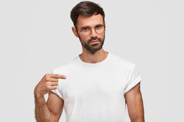 Retrato horizontal de bonito homem com barba por fazer e barba por fazer, vestido com uma camiseta branca casual, aponta para o espaço em branco da cópia para seu projeto, usa óculos. Homem sério vendedor de roupas