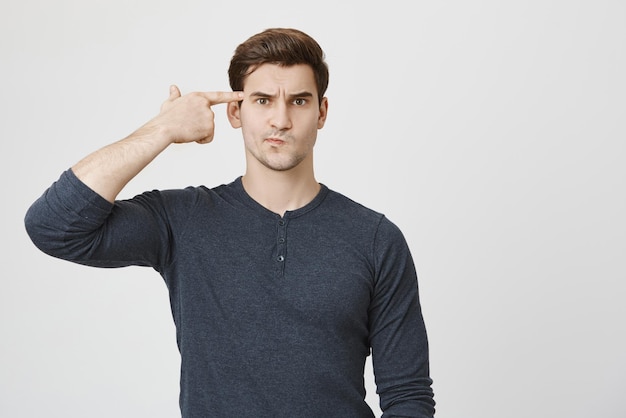 Retrato horizontal de homem caucasiano com barba por fazer e corte de cabelo na moda fazendo beicinho em roupas casuais entediado com o trabalho atirando em si mesmo com gesto de arma de dedo expressões e emoções do rosto humano