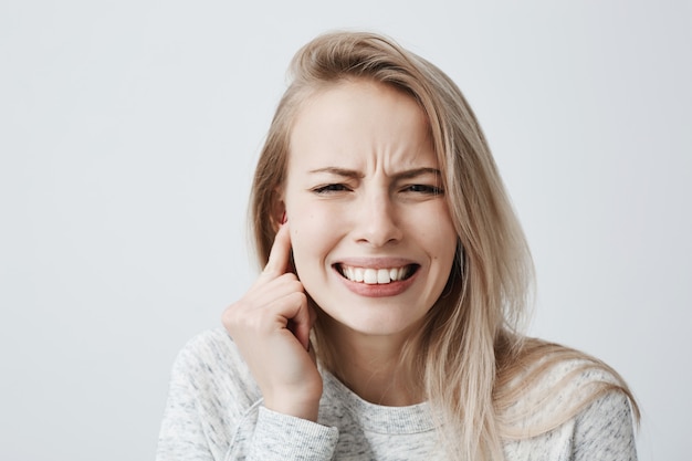 Foto grátis retrato horizontal de loiro caucasiano feminino vestido casualmente tem dor de cabeça depois da festa barulhenta, aperta os dentes e segura a mão atrás da orelha. fêmea jovem irritada expressando emoções negativas.