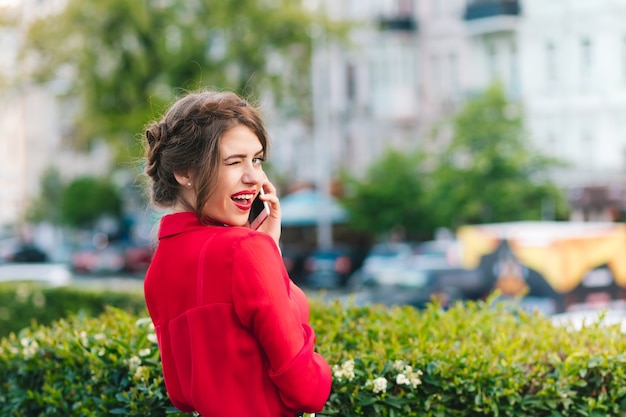 Retrato horizontal de menina bonita em pé no parque. Ela usa blusa vermelha e um belo penteado. Ela está falando ao telefone e faz um sinal com os olhos para a câmera.