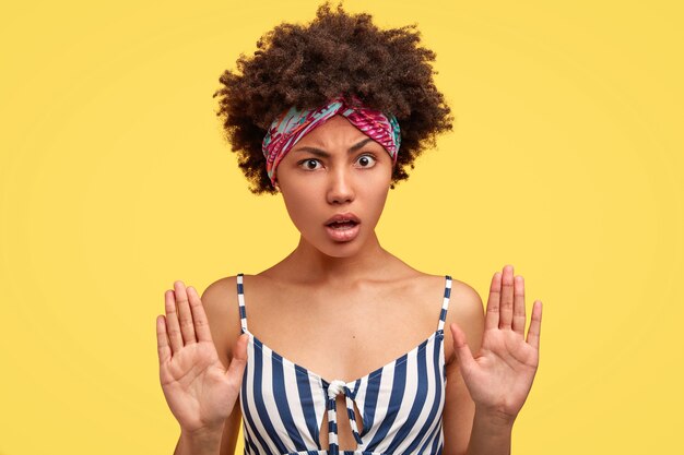 Retrato horizontal de mulher jovem e atraente com penteado afro, pele escura e saudável, expressão facial indignada e descontente, mostra gesto de recusa, isolado sobre a parede amarela. Pare!