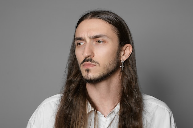 Retrato horizontal de um jovem elegante com a barba por fazer, brinco e penteado comprido e solto, sobrancelhas franzidas, preocupado com o trabalho, posando de camisa branca