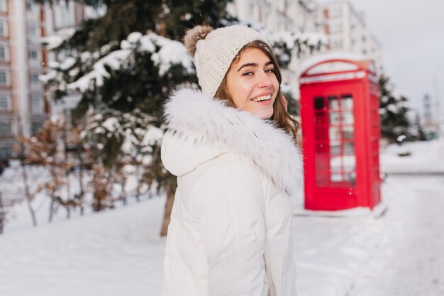 Retrato incrível sorridente jovem inverno andando na rua cheia de neve na manhã ensolarada. Cabine telefônica vermelha, estilo britânico, aproveitando o frio