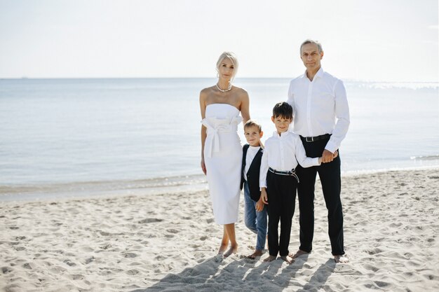 Retrato informal de família na praia de areia no dia ensolarado de verão, de pais e dois filhos vestidos em trajes formais