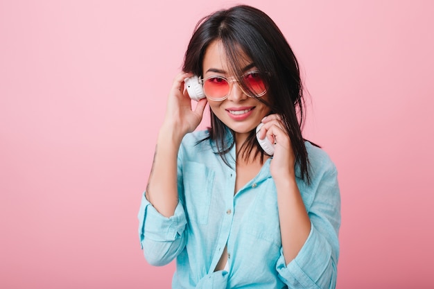 Retrato interior da tímida garota latina em óculos de sol rosa, ouvindo música em grandes fones de ouvido brancos. romântica senhora asiática de cabelos negros na camisa de algodão azul, curtindo a música favorita.