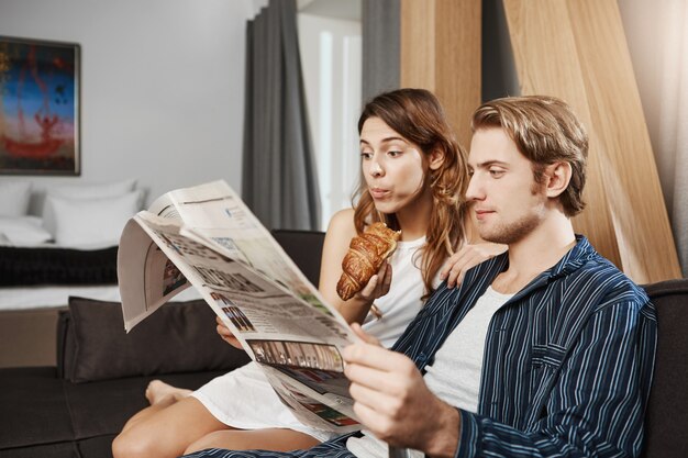 Retrato interior de casal apaixonado lendo jornal no apartamento, sentado no sofá, de pijama. Namorada lê página do horóscopo e come croissant enquanto namorado verifica notícias de negócios
