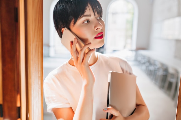 Retrato interior de close-up de mulher jovem ocupada com lábios vermelhos e penteado curto da moda falando ao telefone