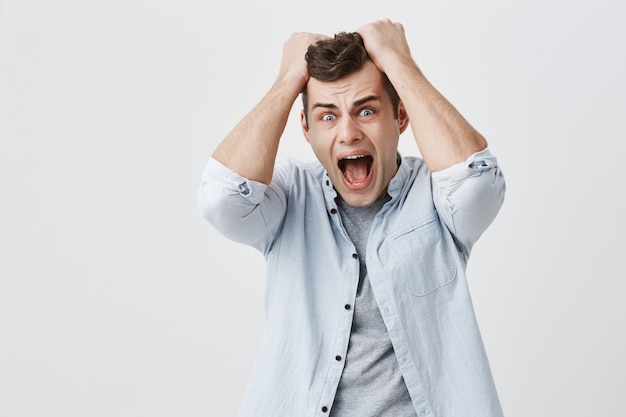 Retrato interior de homem bonito caucasiano estressado, estressado, vestido com camisa azul, segurando as mãos na cabeça e gritando alto em desespero e raiva, furioso com o barulho vindo do apartamento acima dele