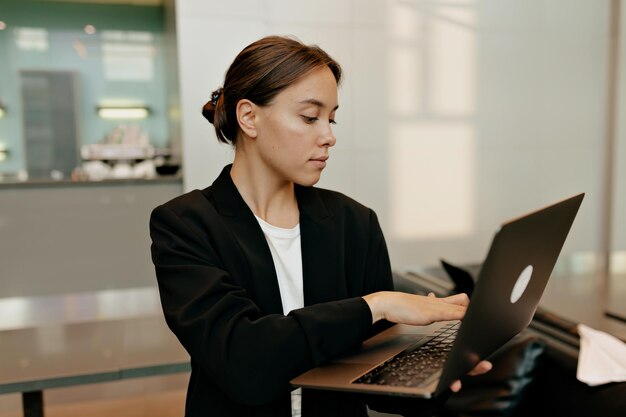 Retrato interior de jovem com cabelo escuro está digitando no laptop e olhando para a tela branca Mulher alegre descansando no escritório durante a pausa para o café