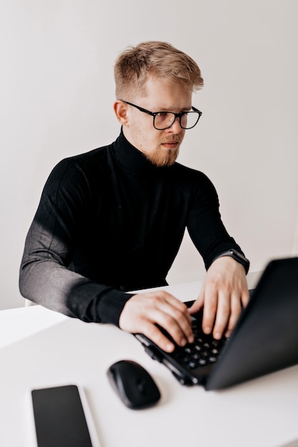 Retrato interior de jovem europeu vestindo pulôver preto e óculos trabalhando com laptop no escritório claro em dia ensolarado.