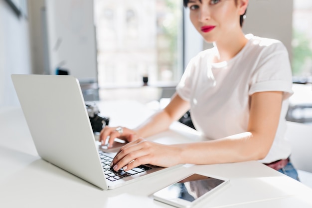 Retrato interior de mulher morena elegante em camisa branca com laptop moderno e smartphone em primeiro plano