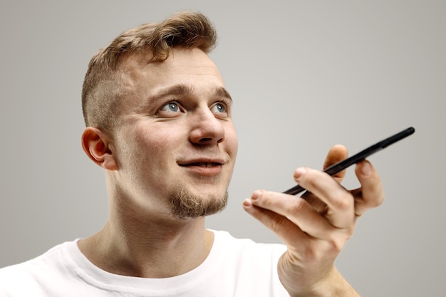 Retrato interior de um jovem atraente isolado em um fundo cinza, segurando o smartphone, usando o controle de voz, sentindo-se feliz e surpreso. Emoções humanas, conceito de expressão facial.