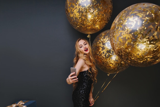 Foto grátis retrato interior de uma deslumbrante garota de cabelos louros fazendo selfie em festivo. mulher espetacular com balões de festa, se divertindo e tirando foto de si mesma.