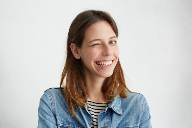 Foto grátis retrato interior de uma linda garota vestida com jaqueta jeans por cima listrado, sorrindo amplamente e piscando misteriosamente, tendo um olhar brincalhão.