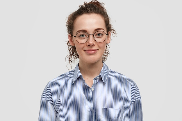 Retrato interior de uma linda mulher sardenta com cabelo escuro e encaracolado, veste a camisa listrada da moda, alegra-se dia de folga, isolado sobre uma parede branca. Mulher cacheada e satisfeita fica sozinha dentro de casa