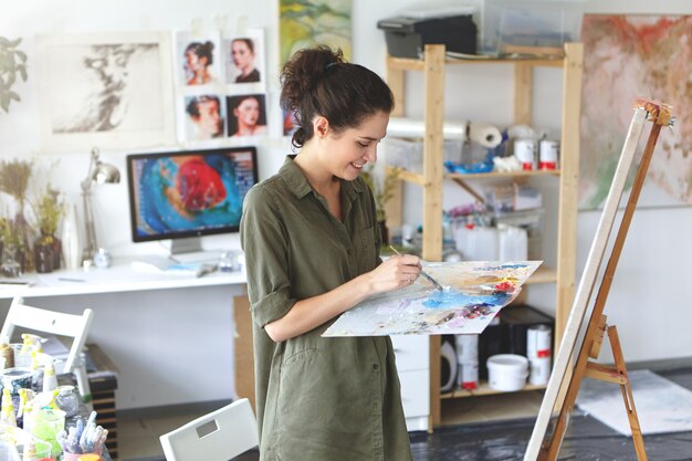 Retrato interior do artista jovem animado feliz na camisa de cor militar, segurando a paleta e o pincel enquanto trabalhava na pintura em sua oficina, em pé na frente do cavalete e sorrindo