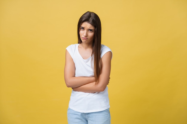 Retrato interno de linda garota em pé com as mãos cruzadas na barriga, sentindo-se estranho ou sofrendo de dor ao olhar para o lado, em pé contra um fundo amarelo. Mulher tem dor de estômago.