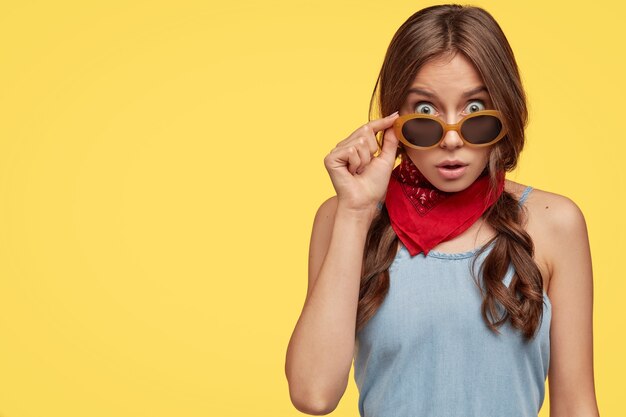 Retrato interno de menina chocada vê algo inacreditável, olhando através de óculos escuros