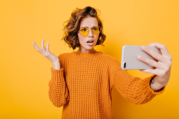 Retrato interno de mulher decepcionada de cabelo curto de óculos fazendo selfie no estúdio