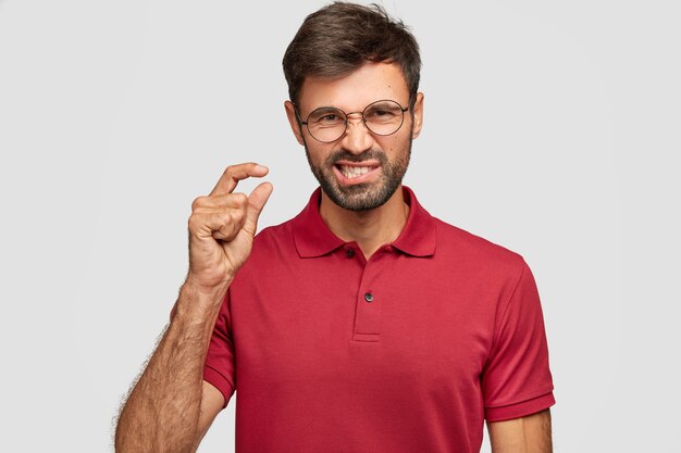 Retrato interno de um jovem bonito descontente com a barba por fazer franze a testa em insatisfação, gesticula com a mão, mostra algo muito pequeno, vestido com uma camiseta vermelha, isolado sobre uma parede branca