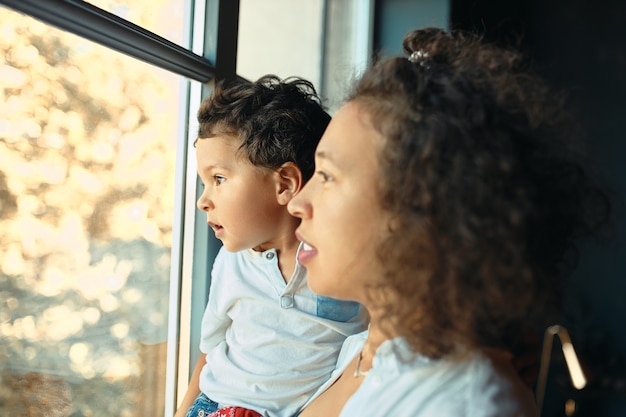 Retrato interno de uma jovem mãe latina feliz ficando em casa em frente à janela com o filho pré-escolar nos braços, olhando para fora através do vidro