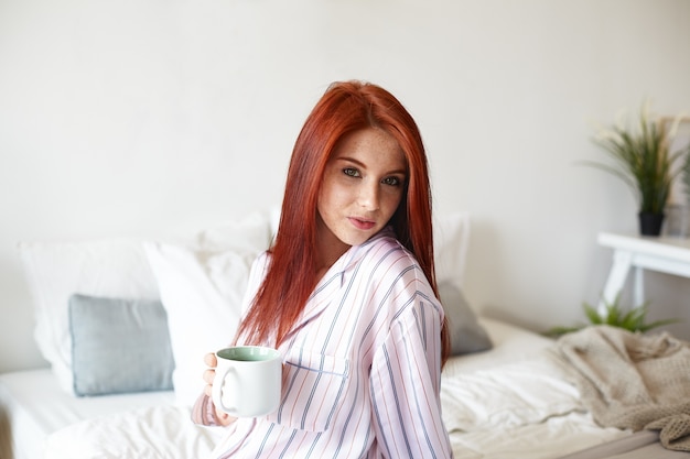 Retrato interno de uma linda jovem ruiva caucasiana com roupa de noite listrada, começando um novo dia, desfrutando do café da manhã, sentada no quarto com uma caneca grande e olhando