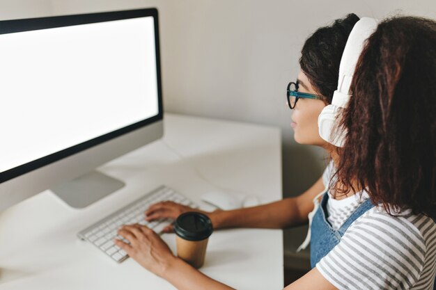 Retrato interno de uma mulher de cabelos castanhos usando óculos e fones de ouvido, trabalhando no computador e bebendo café