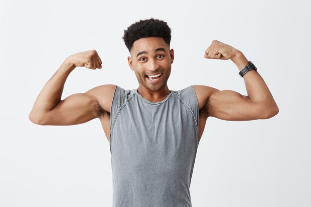 Retrato isolado de alegre alegre atraente atlético homem de pele escura com penteado afro na camisa cinza desportiva, mostrando os músculos grandes, olhando na câmera com expressão feliz e relaxada.