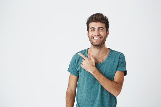 Retrato isolado de feliz caucasiano atraente em casual camiseta azul, com penteado na moda, sorrindo e apontando para a parede em branco. Copie o espaço.