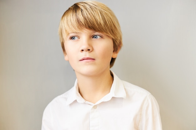 Retrato isolado de um menino branco espantado com franjas e olhos azuis desviando o olhar com uma expressão pensativa misteriosa, imerso em pensamentos, pensando, tendo uma ideia ou fazendo um plano, posando para uma parede em branco