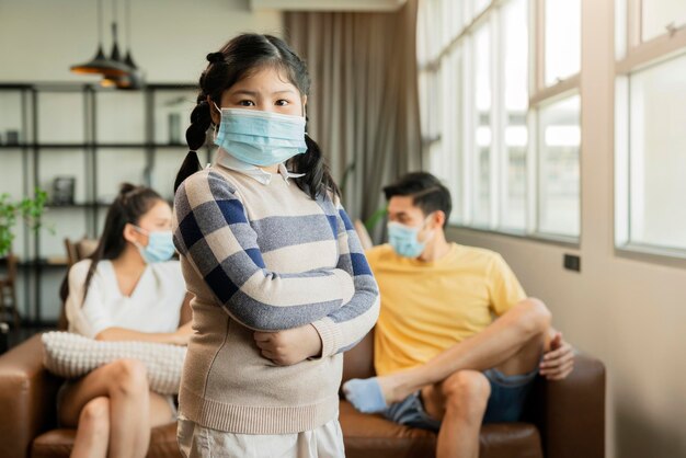Retrato jovem menina asiática usando máscara cirúrgica facial ficar forte posando confiante em quarentena distanciamento social em casa com a família na sala de estar