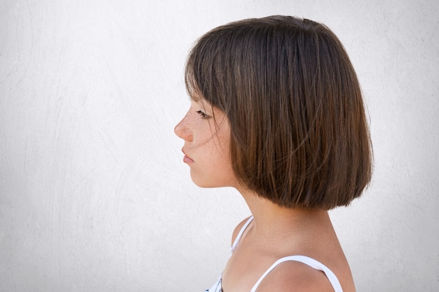 Foto grátis retrato lateral da menina sardenta adorável que olha na distância ao ter a expressão sonhadora isolada no muro de cimento branco. menina com cabelo escuro e curto, de pé para o lado com olhar sério