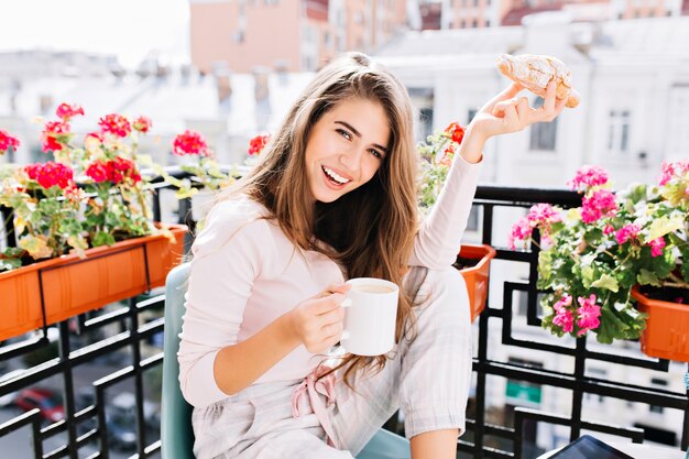 Retrato linda garota com cabelo comprido, tomando café da manhã na varanda cercam flores pela manhã na cidade. Ela segura uma xícara, croissant, se divertindo.