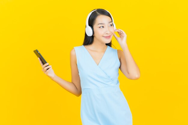 Retrato linda jovem asiática com fone de ouvido e telefone inteligente para ouvir música na parede amarela