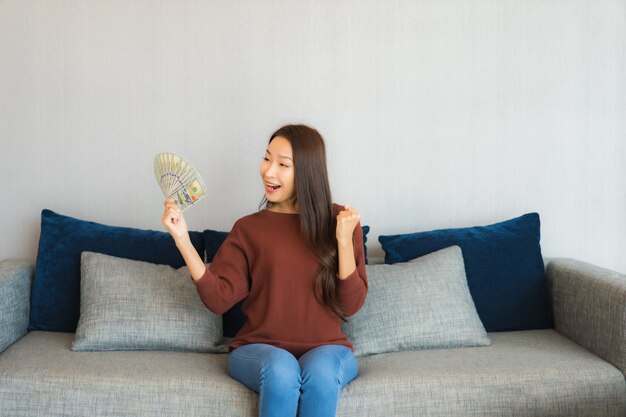 Retrato linda jovem asiática mostrar dinheiro e dinheiro no sofá no interior da sala de estar