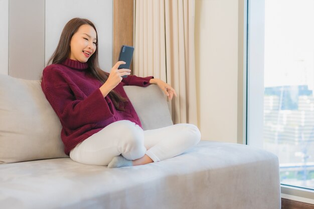 Retrato linda jovem asiática usando celular inteligente no sofá no interior da sala de estar