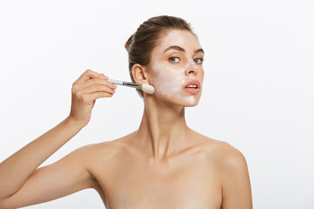 Retrato linda mulher nua aplica máscara de argila branca cosmética com pincel isolado nas costas brancas