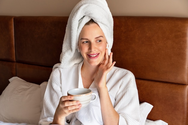 Retrato matinal de uma linda mulher relaxando na cama após o banho, usando roupão de banho e toalha na cabeça, bebendo um chá saboroso