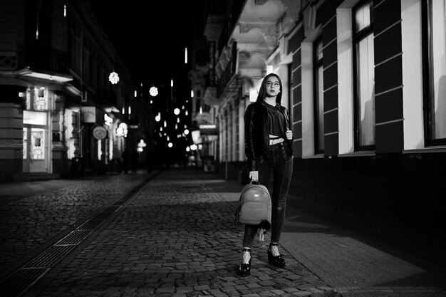Retrato noturno do modelo de menina vestindo óculos jeans e jaqueta de couro com mochila contra as luzes das ruas da cidade