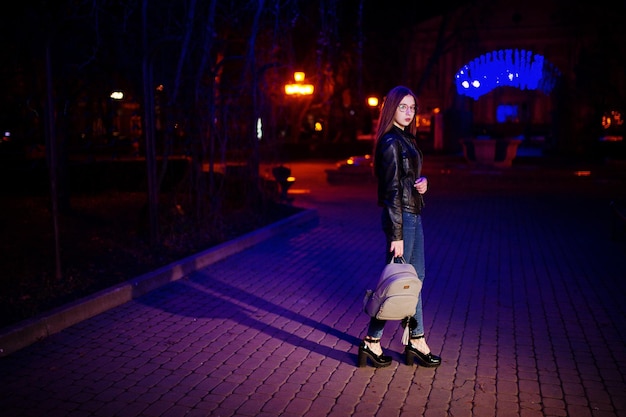 Retrato noturno do modelo de menina vestindo óculos jeans e jaqueta de couro com mochila nas mãos contra guirlanda de luzes azuis da rua da cidade