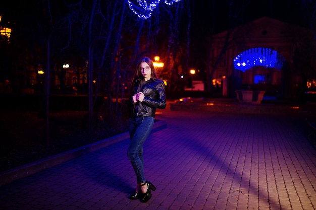 Retrato noturno do modelo de menina vestindo óculos jeans e jaqueta de couro contra luzes azuis guirlanda de rua da cidade