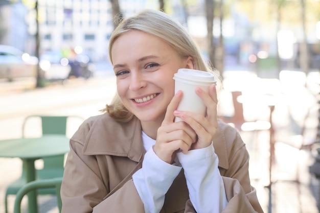 Foto grátis retrato próximo de mulher loira sorridente segurando uma xícara de café sentada em um café de rua ao ar livre