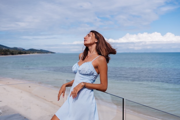 Retrato romântico de mulher com vestido azul claro sozinha em uma praia tropical, dia de sol, pele morena bronzeada