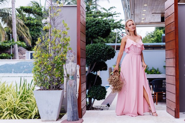 Retrato romântico de mulher com vestido de noite rosa segurando flores silvestres do lado de fora da luxuosa villa tropical. Linda mulher com buquê
