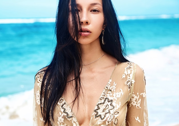 Retrato sensual do modelo de mulher bonita caucasiano com cabelos longos escuros na blusa bege posando na praia de verão com areia branca no fundo do céu e oceano azul